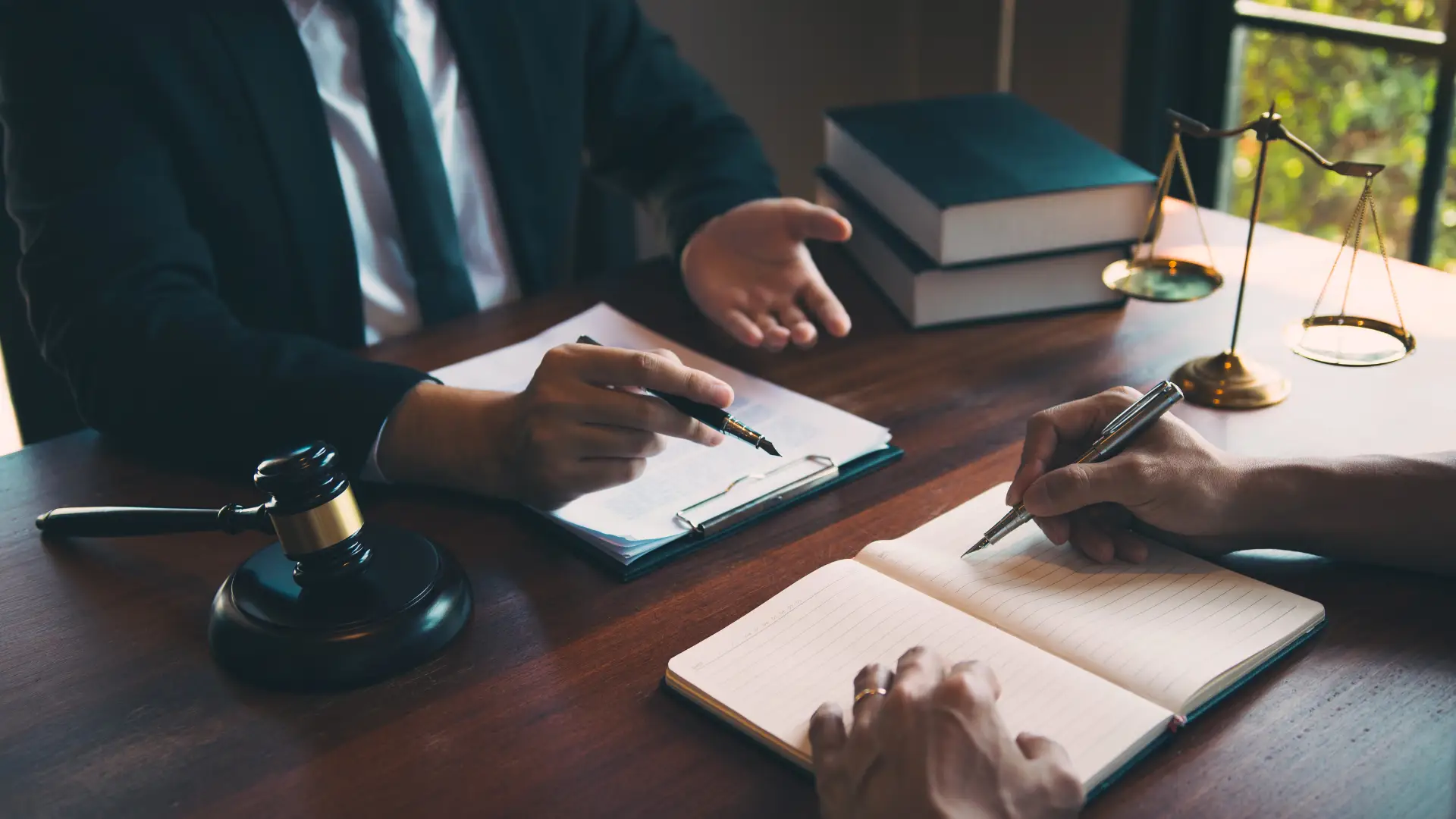 Two individuals sitting at a desk with legal books and documents, engaging in a discussion, possibly in a law office setting.